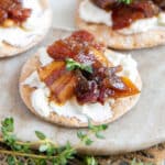 A marble board bearing crackers topped with cream cheese, bacon jam and thyme leaves