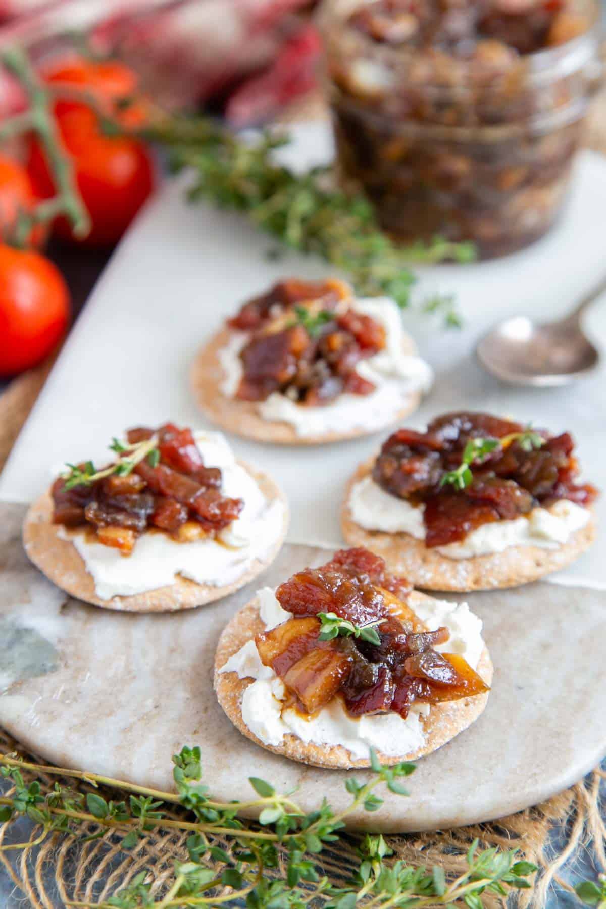 A marble board bearing crackers topped with cream cheese, bacon jam and thyme leaves
