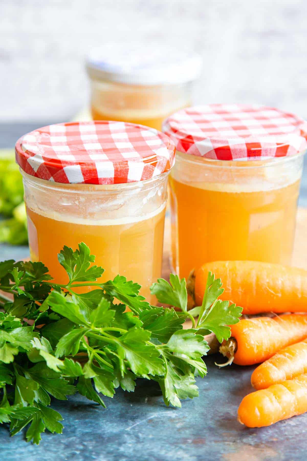 Jars of classic chicken stock surrounded by fresh parsley and carrots.