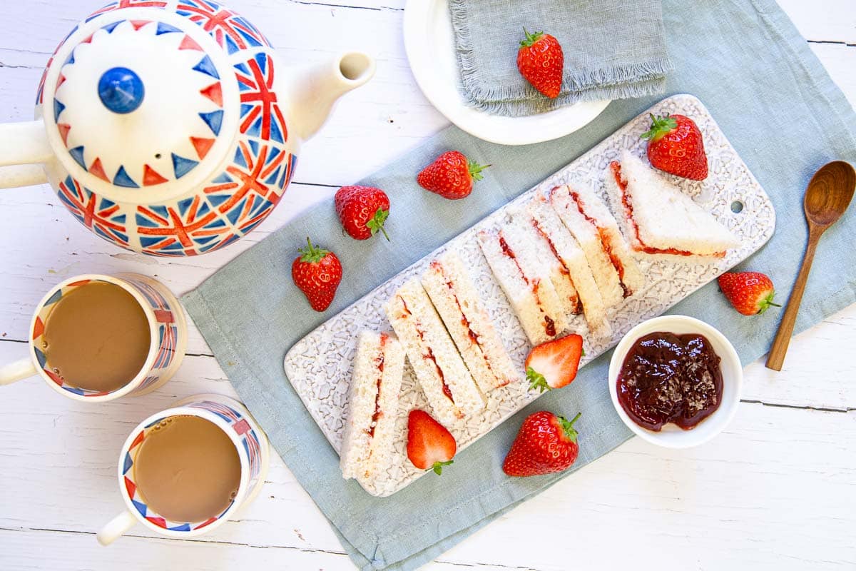 The perfect sandwich tea - strawberry jam sandwiches seen from above, scattered with fresh strawberries and accompanied by a tea pot, two mugs of tea and a dish of jam.