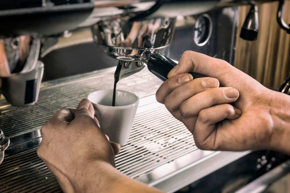 A close up of an espresso machine; coffee is filling an espresso cup. The barista is holding both the cup and the filter.