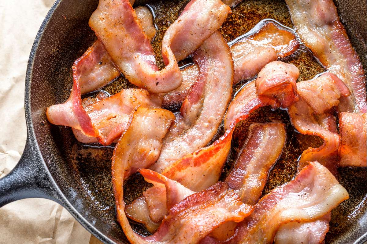 A cast iron frying pan full of rashers of streaky bacon being cooked.