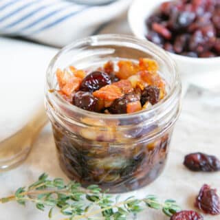 A pot of cranberry and bacon jam on a marble board with a sprig of thyme