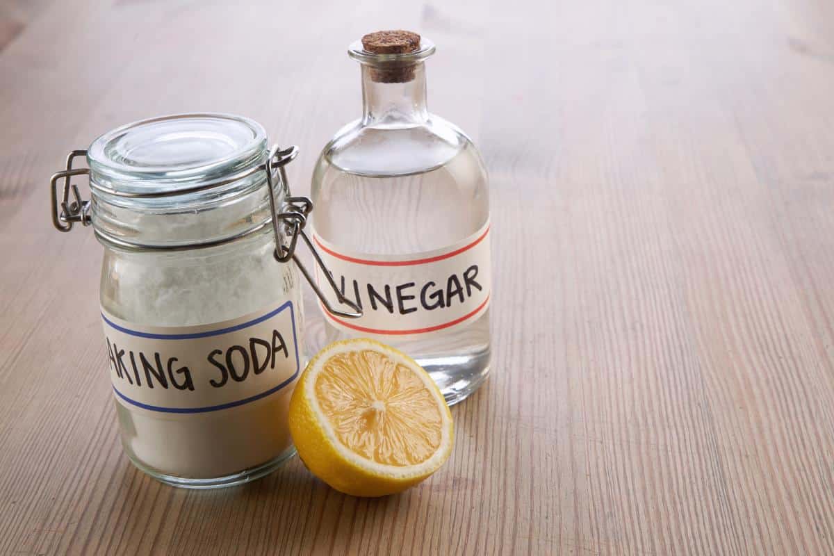 A bottle of vinegar, a clip top jar of baking soda and half a lemon, all on a wooden surface.