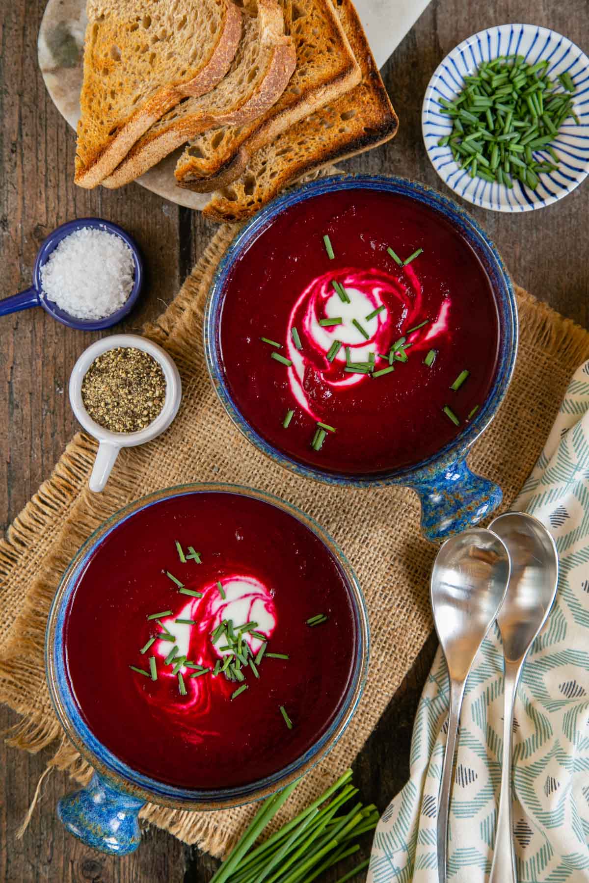 Two dishes of ruby red beetroot soup shown from above, swirled with sour cream and scattered with chives.