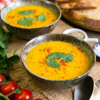 A table spread for lunch with bowls of golden carrot and coriander soup, crusty bread and tomatoes.