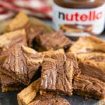 A pile of delicious nutella swirled chocolate hazelnut blondies with a jar of Nutella behind.
