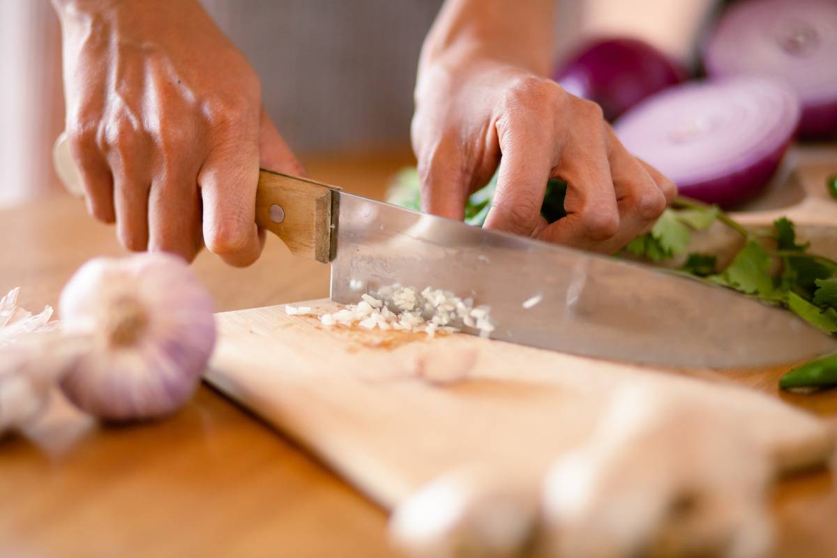 A large wooden handled knife is being used to finely chop garlic, on a wood board. More garlic and other vegetables surround.