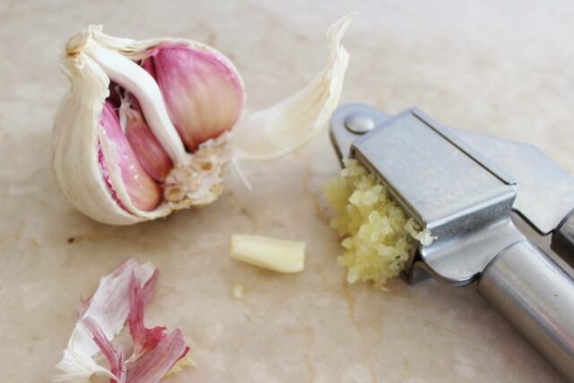 A garlic press that has processed some garlic, and half a head of garlic.