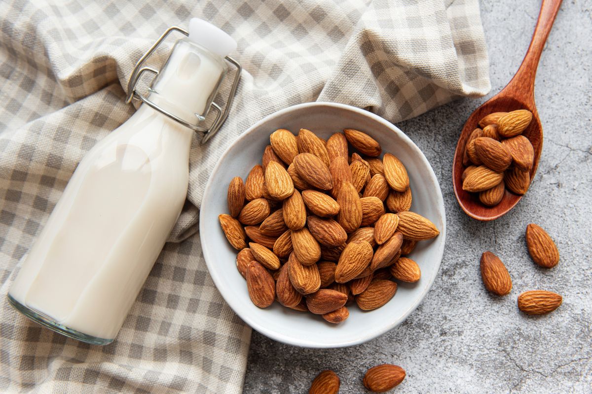 Almonds in a bowl and wooden spoon, next to a clip-top bottle of almond milk, sitting on a white and grey gingham cloth.
