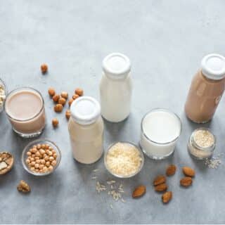 Bottles and glasses of plant milk and respective seeds, nuts and vegetables, on a pale grey background.