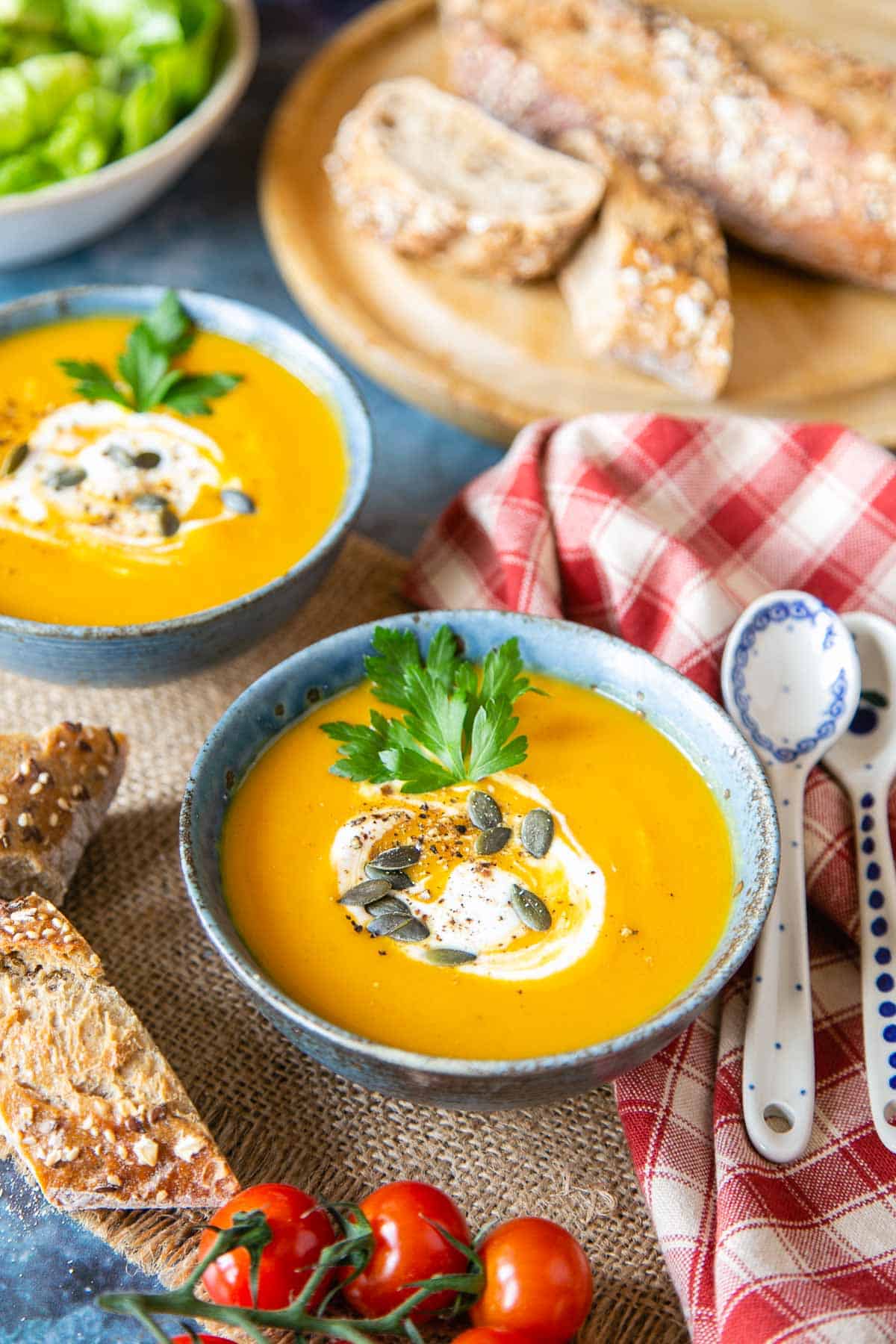 a table set with two bowls of golden sweet potato and carrot soup, fresh bread, a green salad and vine tomatoes
