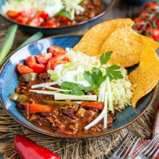 A blue glazed bowl full of deep, dark beef mince chilli with toppings and tacos.