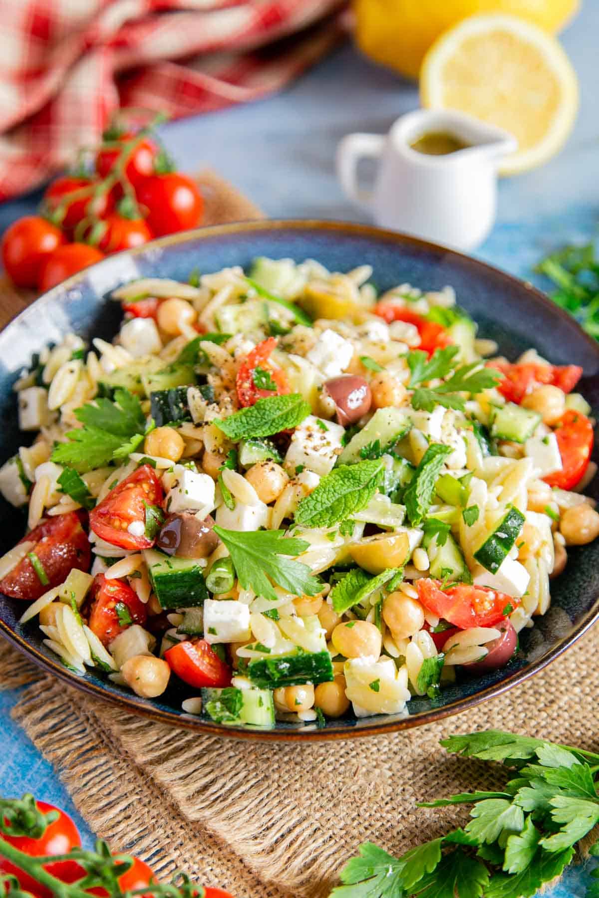 A dish of Greek orzo salad surrounded with fresh herbs and tomatoes.