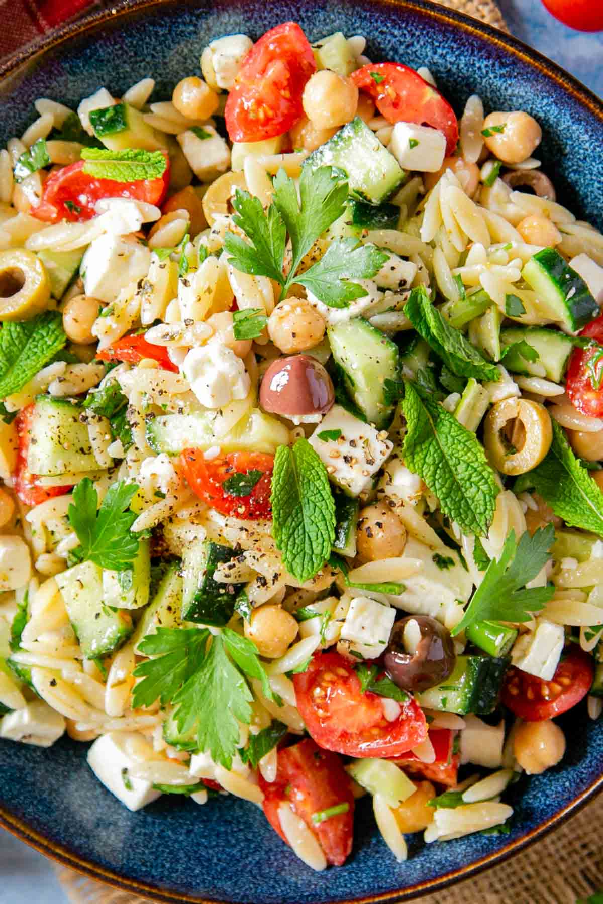 A bowl of Greek orzo salad in close up, full of feta, fresh vegetables, chickpeas and herbs.