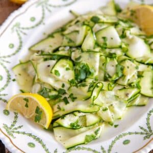 A plate of courgette salad, light and bright courgette ribbons in lemon dressing with lemon wedges.