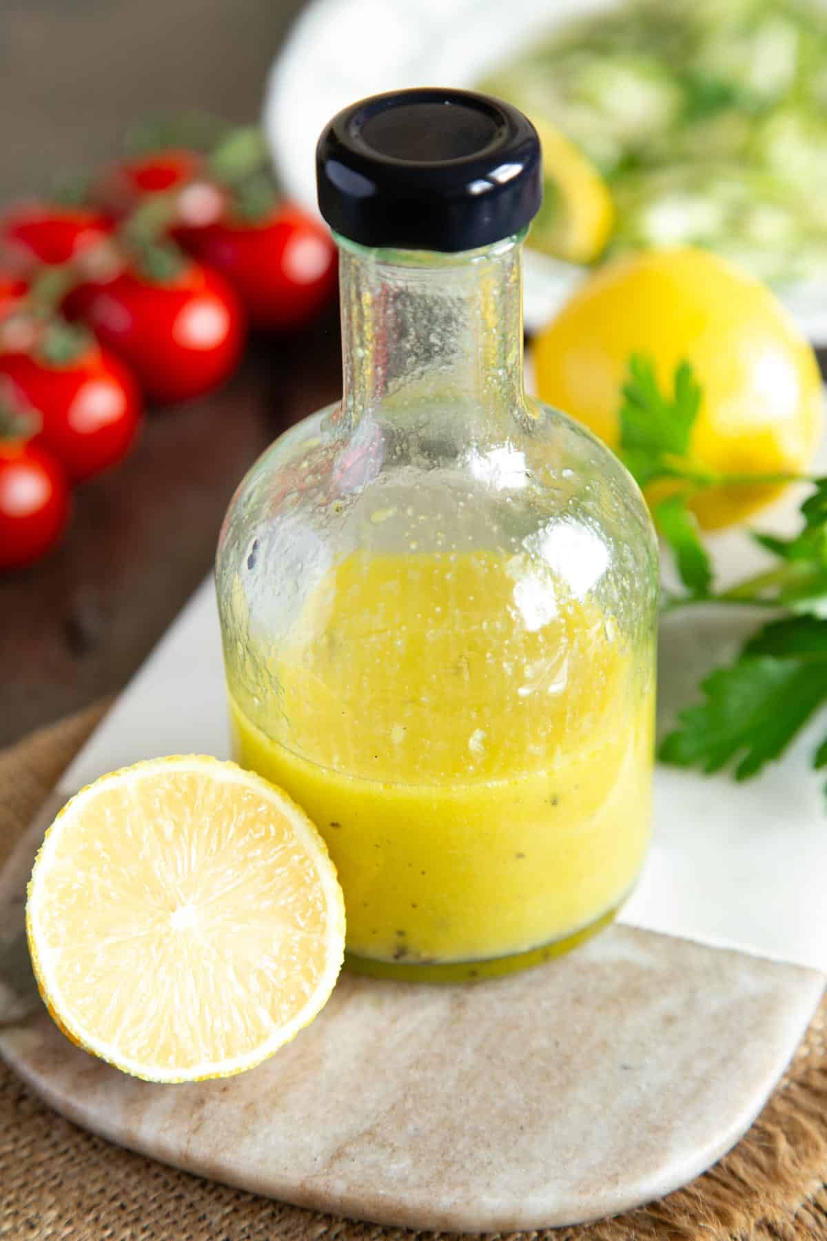 A small bottle of lemon dressing presented on a marble board with lemons and herbs.