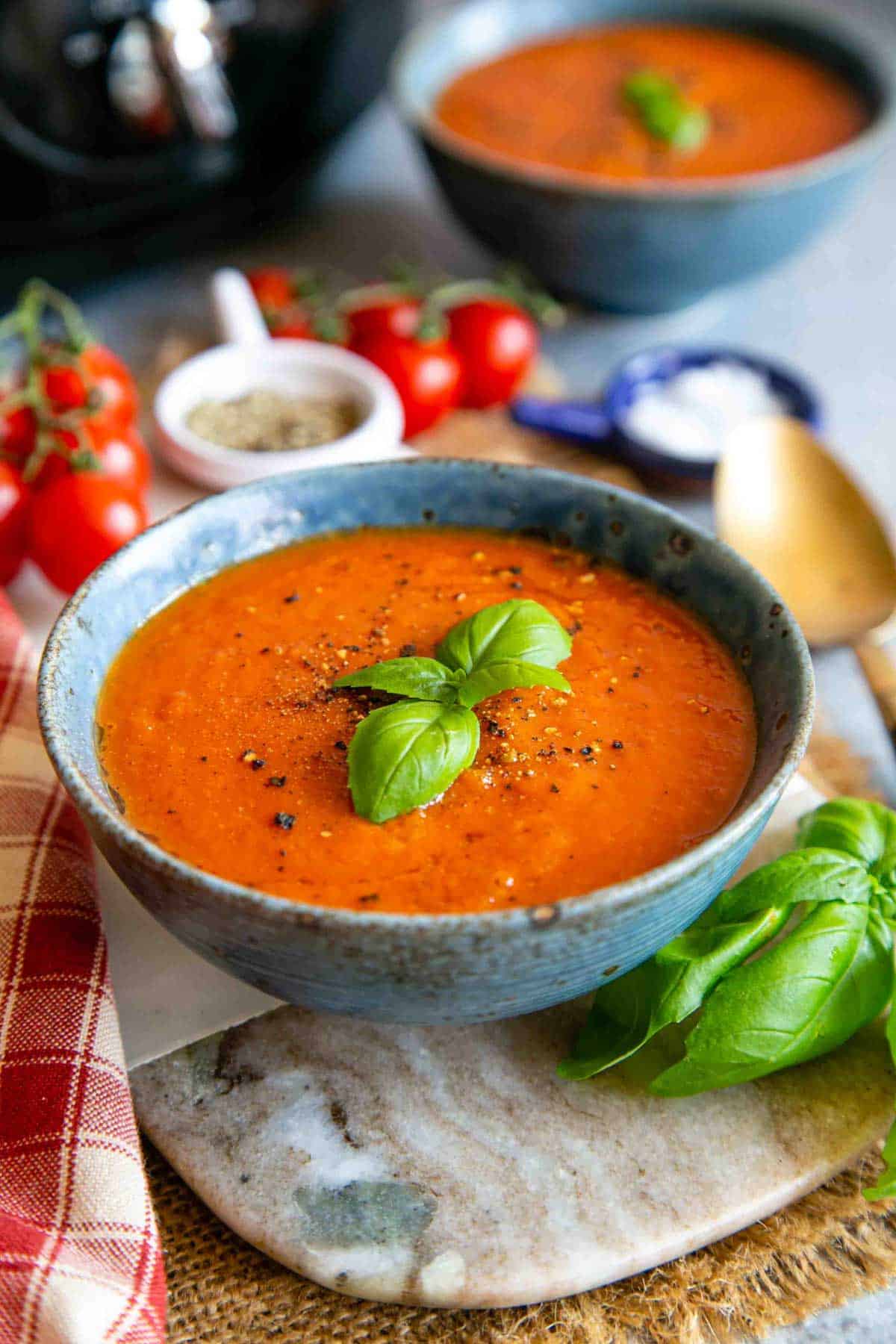 A bowl of delicious slow cooker tomato soup, garnished with a sprig of basil, a string of vine tomatoes in the background.