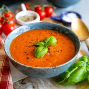 Vibrant red slow cooker tomato soup in a rustic blue bowl, topped with a sprig of green basil.