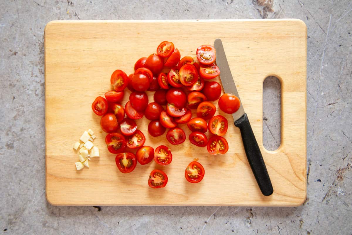 The cut cherry tomatoes on a cutting board with chopped garlic.
