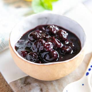 Blueberry compote, deep purple and full of fruit, served in a cream coloured bowl on a pale marble board, next to a halved lemon and a sprig of mint.