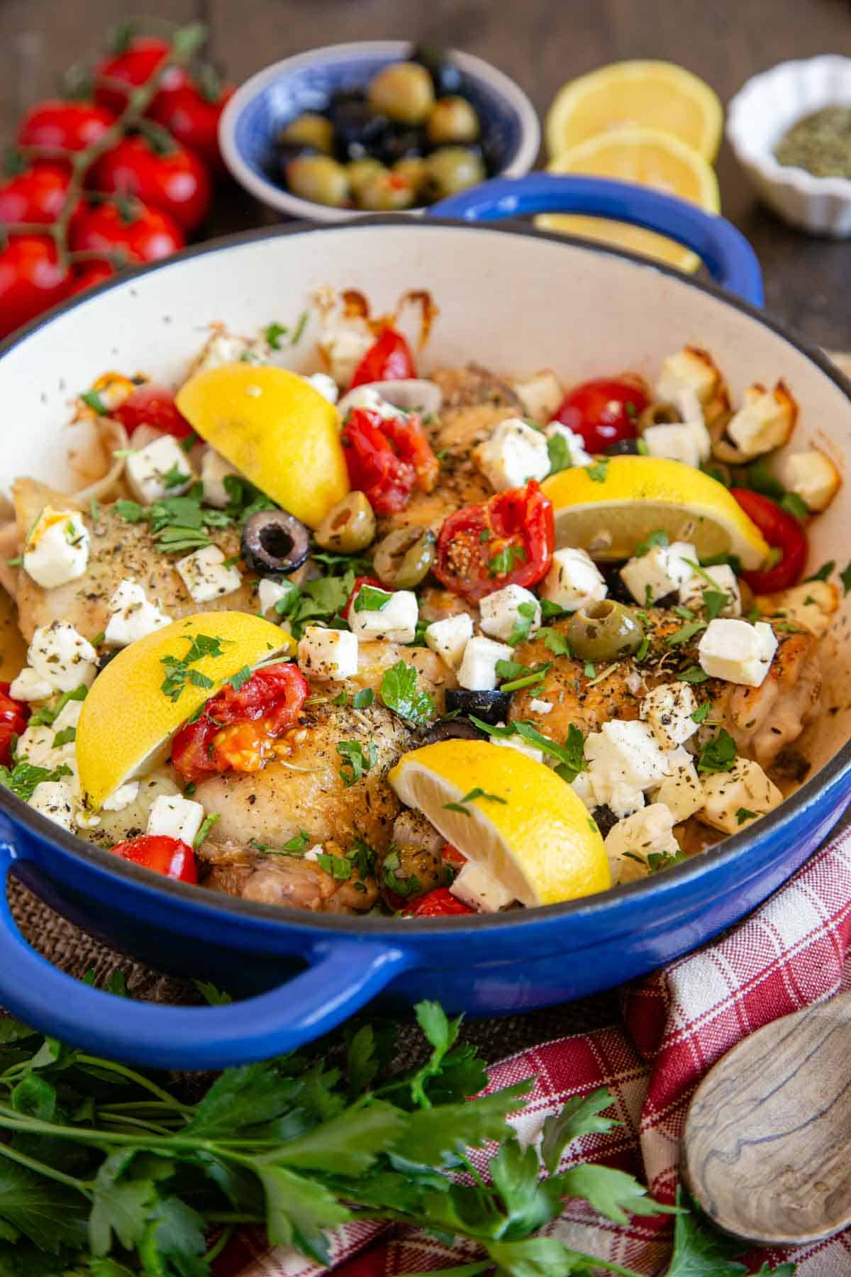 A blue pan of Greek chicken against a red and white checked cloth, surrounded by fresh parsley, tomatoes and lemons.