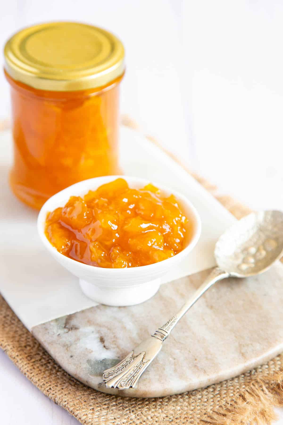 Golden mango jam in a jar and in a dainty serving dish, with a silver spoon alongside.