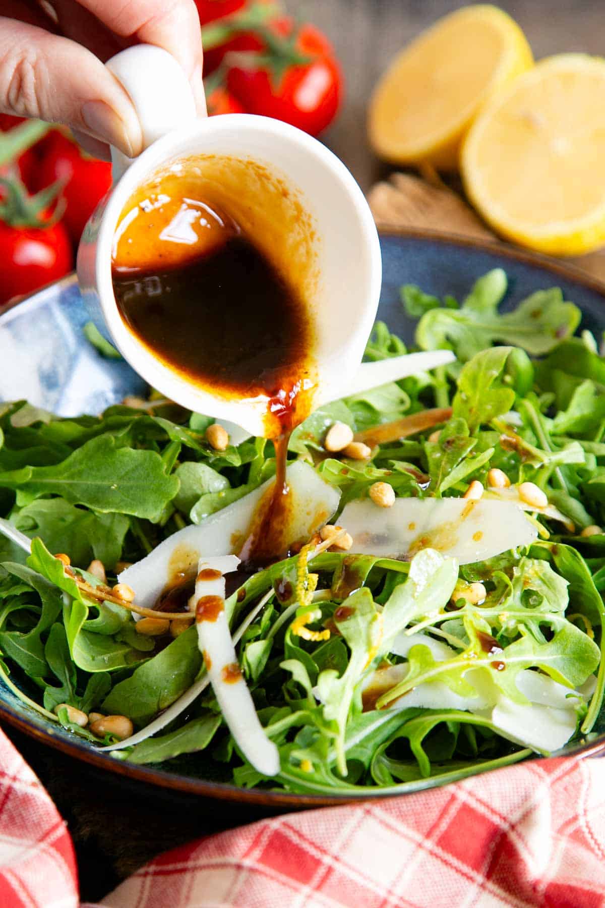 Pouring balsamic dressing from a small jug over a green salad with parmesan shavings.