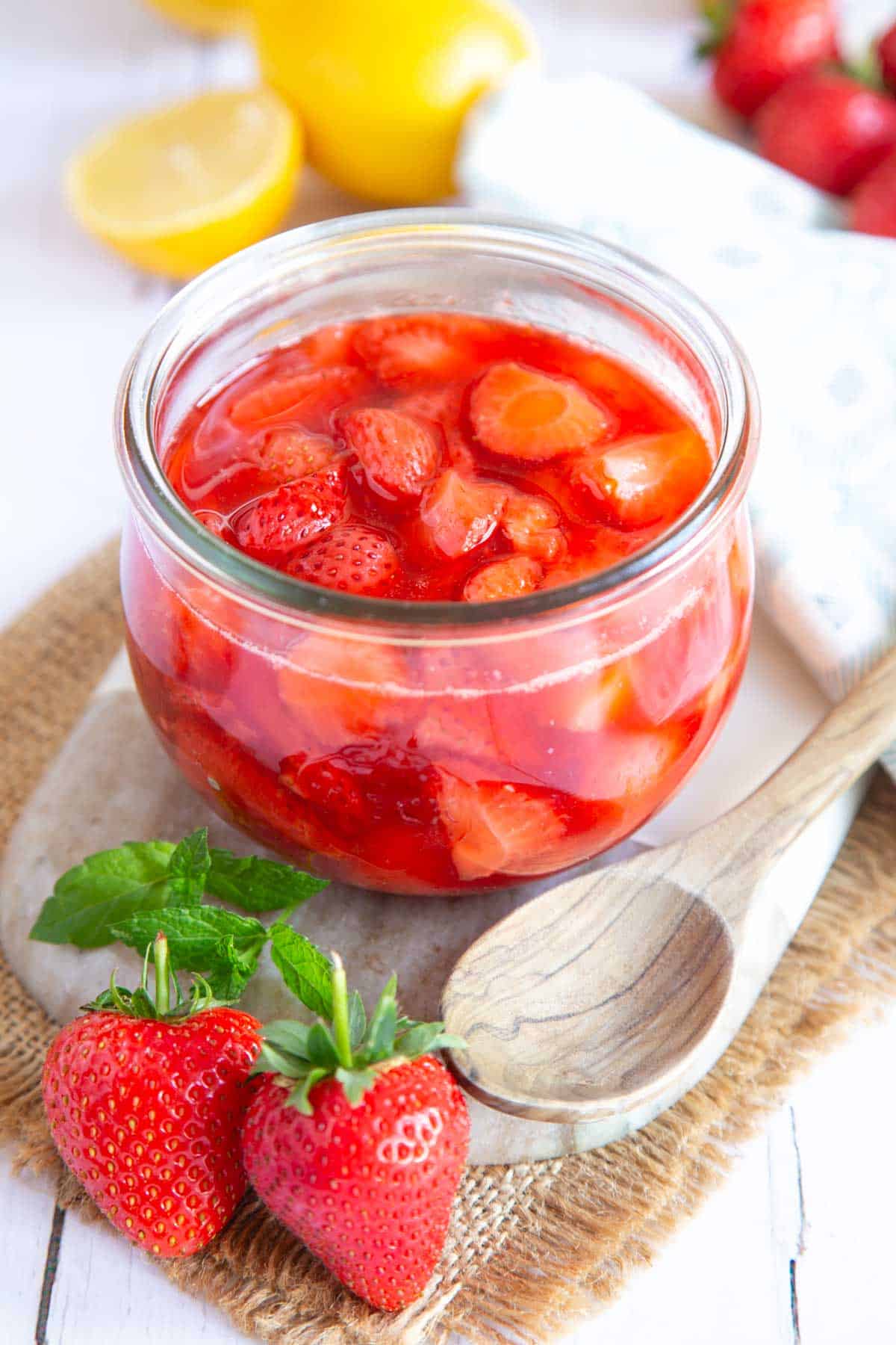 Strawberry compote in a glass jar on a marble serving board, with fresh strawberries and a cut lemon.