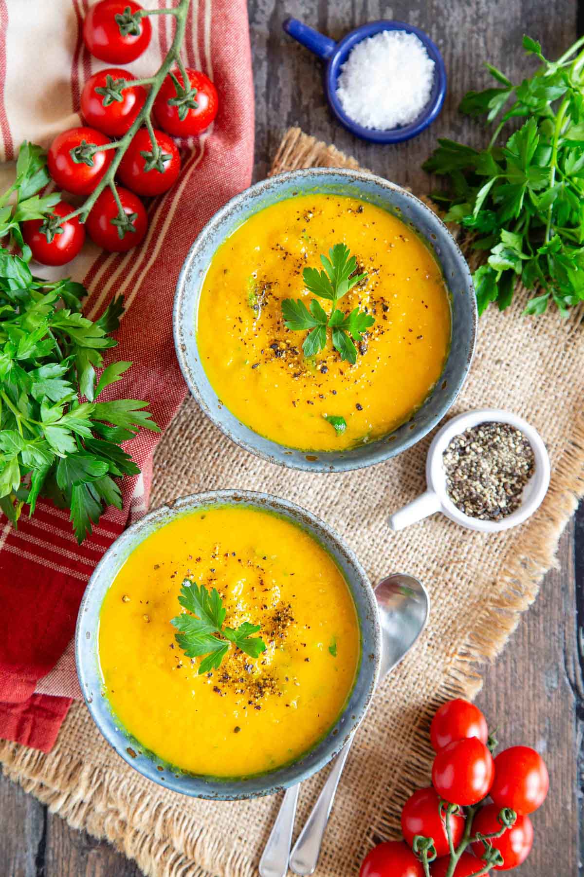 Shown from above, two bowls of carrot and swede soup garnished with extra parsley, on a table surrounded with cherry tomatoes and herbs.