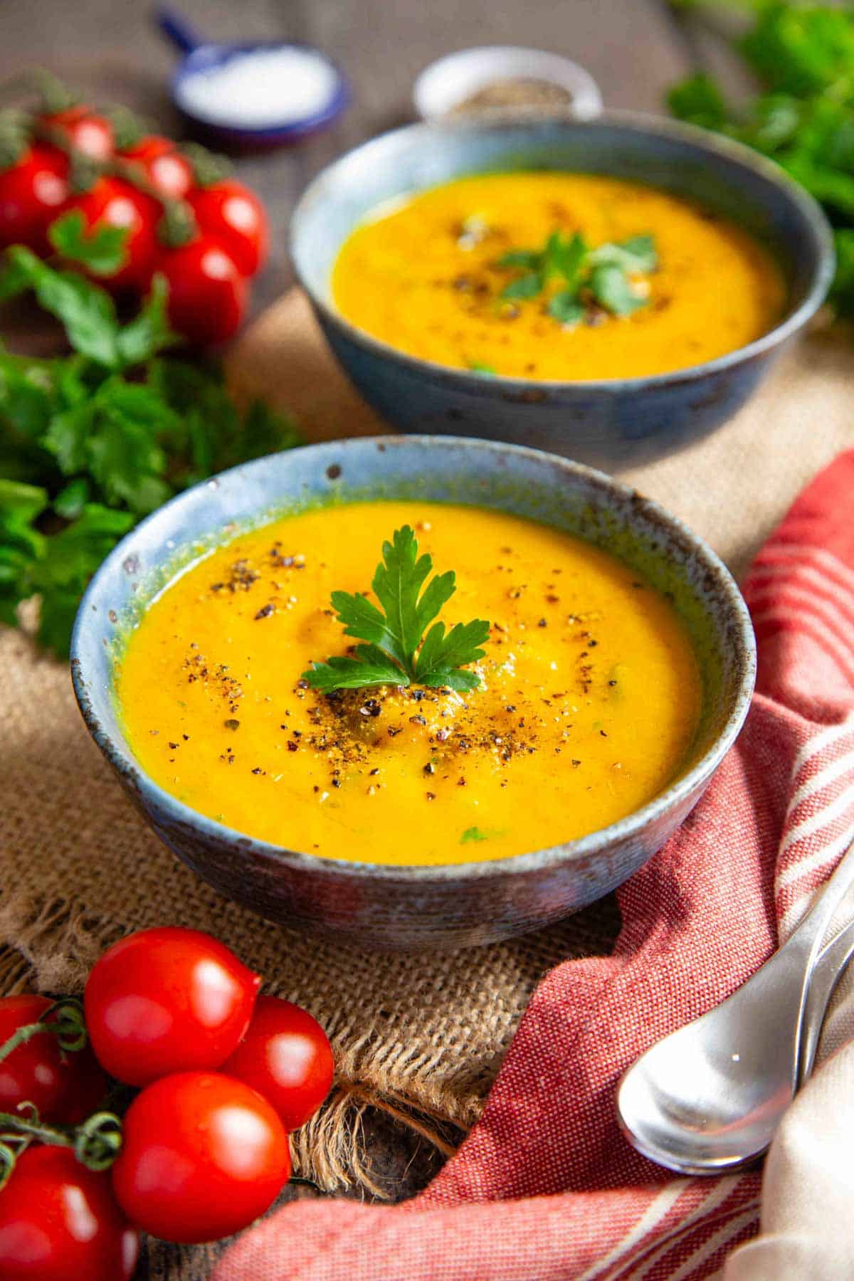 Thick, golden carrot and swede soup in earthenware bowls on a table surrounded by cherry tomatoes and parsley.