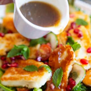 Pouring pomegranate dressing over a salad of grilled halloumi, fresh vegetables, mint and pomegranate seeds.