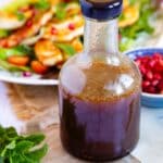 A bottle of pomegrate dressing, with sprigs of mint and a dish of pomegranate, a halloumi salad dish in the background.