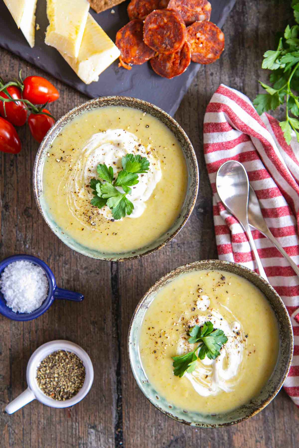 Two bowls of slow cooker leek and potato soup from above, a board with cheese, charcuterie and tomatoes on the side.