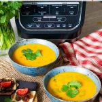 Two bowls of carrot and coriander soup next to the soup maker, with crackers and cheese on the side.
