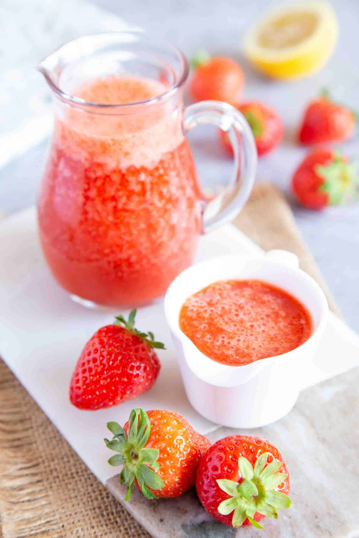 Two jugs of strawberry puree and fresh strawberries on a table