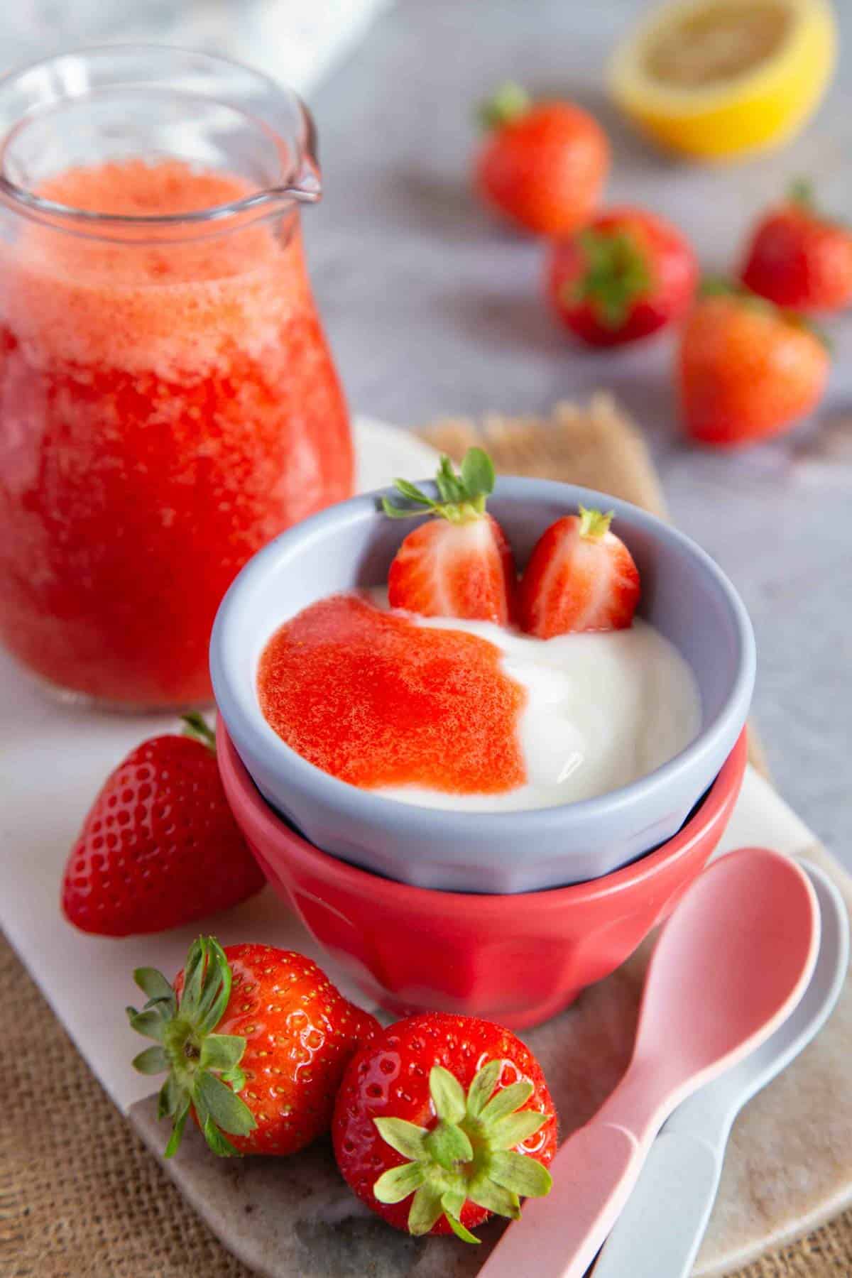 a bowl of yoghurt topped with strawberry puree fresh strawberries and a jug of puree are by the bowl