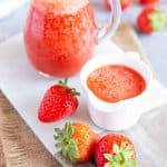 Two jugs of strawberry puree and fresh strawberries on a table
