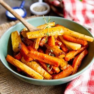 a spoon lifting out a serving of the shiny glazed honey roasted carrots and parsnips from a serving bowl