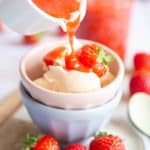 Strawberry coulis is being poured from a jug over a bowl of vanilla ice cream