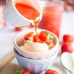 Strawberry coulis is being poured from a jug over a bowl of vanilla ice cream