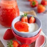 a bowl of yoghurt topped with strawberry puree fresh strawberries and a jug of puree are by the bowl