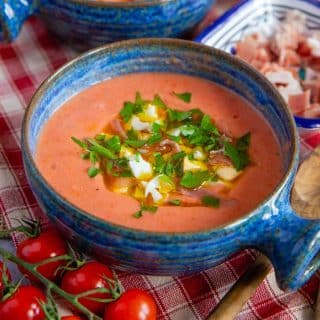 a blue soup bowl full of salmon pink salmorejo garnished with chopped hard boiled egg ham parsley and olive oil