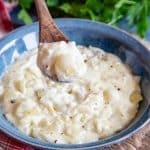 A wooden spoon of onion sauce held in the foreground above a bowl holding more sauce