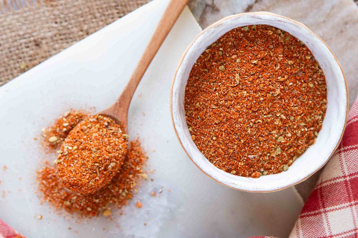 Fajita seasoning red mixture contrasting in a small white bowl with a wooden spoon of mixture to the left