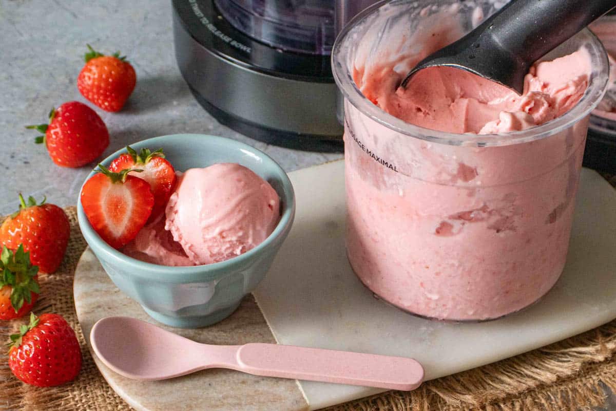 Close up of strawberry ice cream in a pot and bowl with more fruit alongside