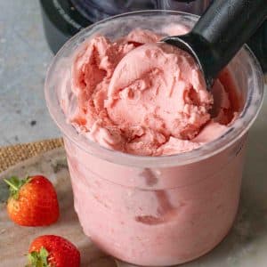 A close up of a pot of pale pink strawberry ice cream with two strawberries in the foreground