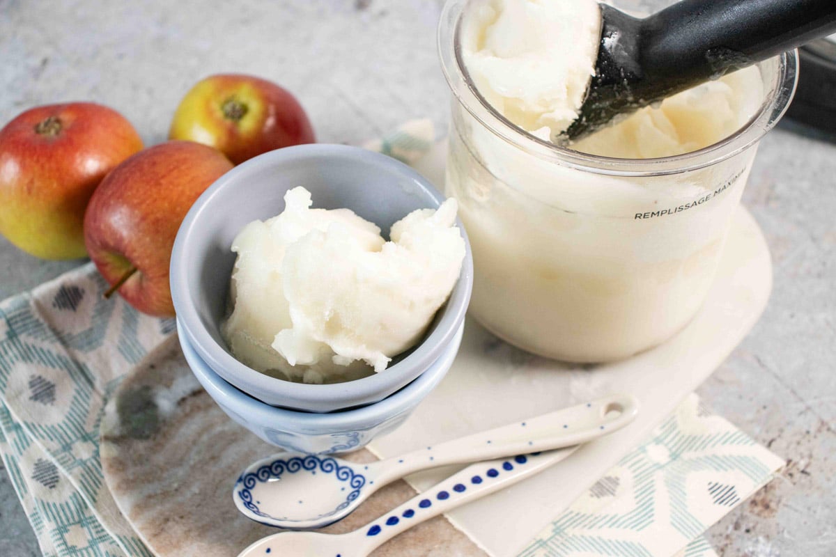 A scoop of tasty pale apple sorbet in a small blue bowl next to the Ninja Creami pot all on a marble board